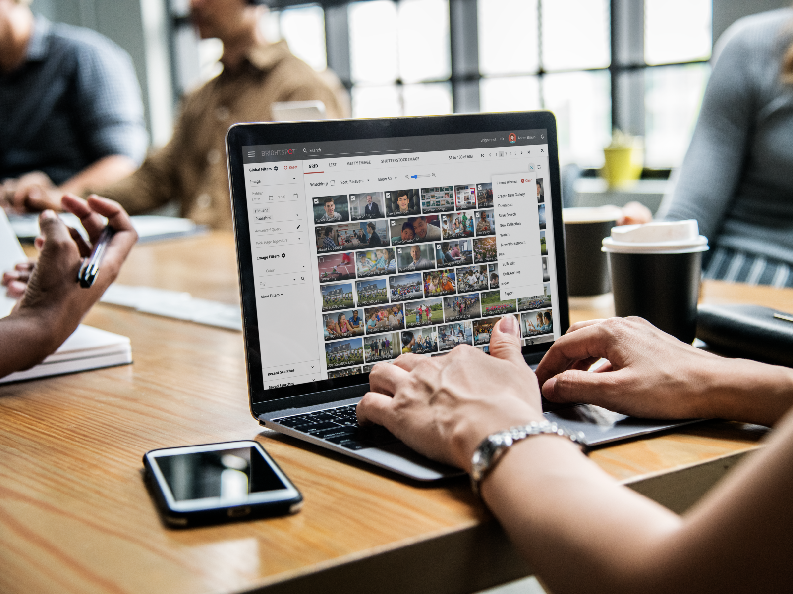 A person using a laptop with an image library on the screen, surrounded by colleagues modern workspace to discuss work process