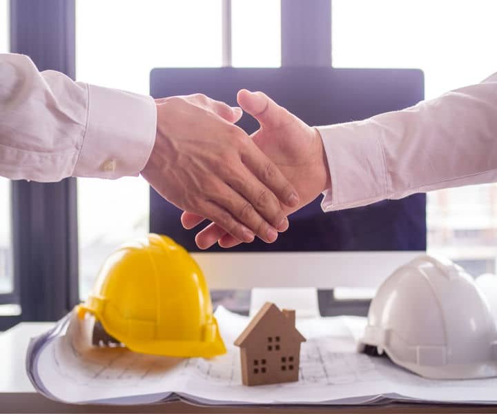 Agreement over a construction plan, with architectural or engineered hats and a small house model on the table in a bright office setting.