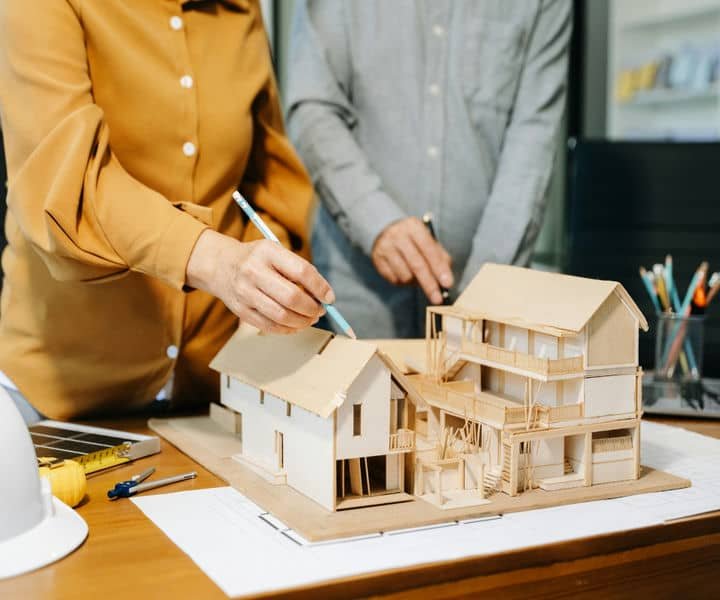 Two architects examine a detailed architectural model of houses, one drawing on it while the other gestures, in a design studio.