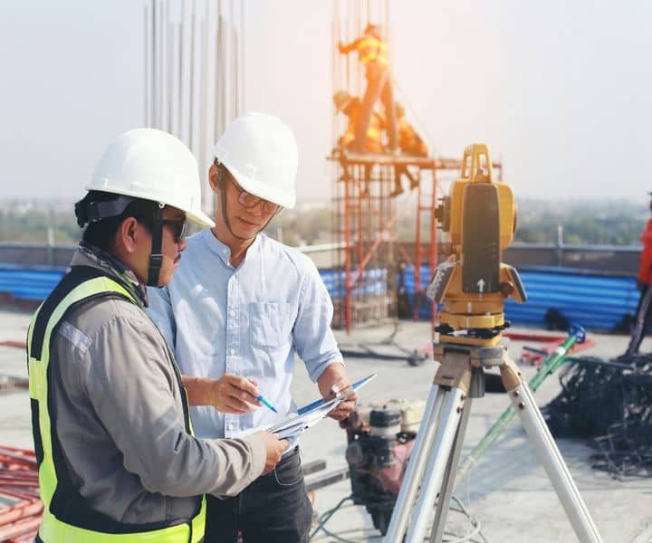Two engineers at construction site review blueprints next to a surveying instrument, with workers constructing a framework.