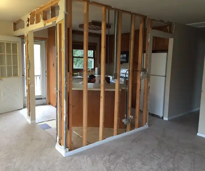 An open-concept room under renovation, featuring exposed wooden studs dividing a kitchen and living area, with unfinished walls.