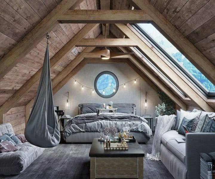 Welcoming attic bedroom with a grey bed, hammocks, round window, and wooden beams, adorned with string lights and stylish decor.