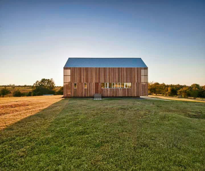 Modern wooden barn with a metal roof, situated on grassy land under a clear sky, showcasing minimalist architecture and open space.