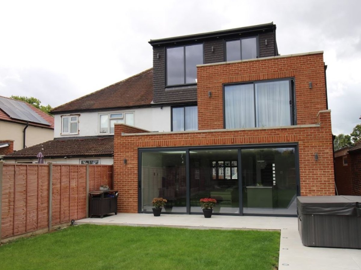 A modern double-story brick house extension with large glass patio doors, surrounded by a garden, featuring a hot tub and neatly trimmed grass.
