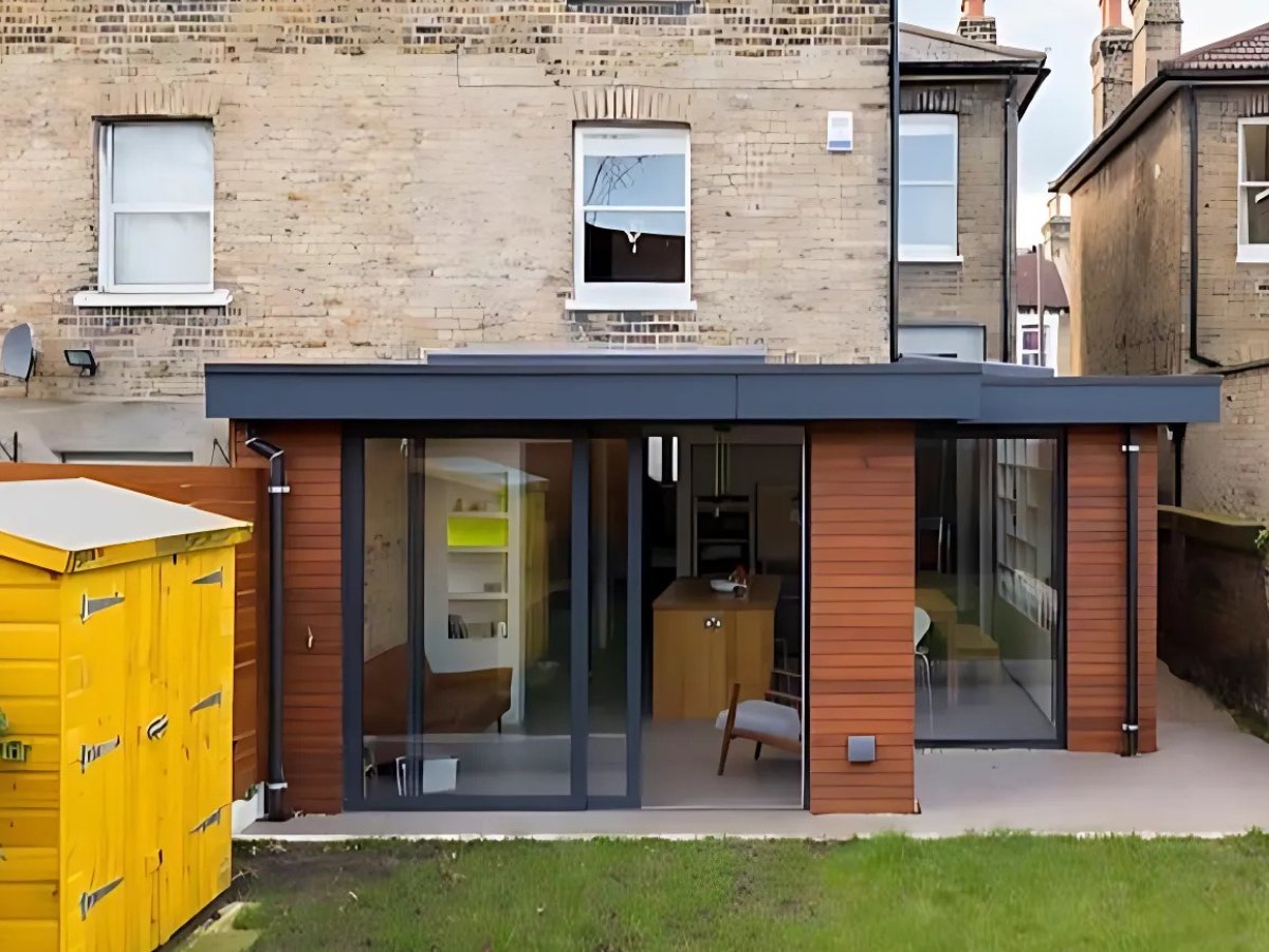 Modern single storey side extension of a brick house featuring large glass doors, wooden accents, and a yellow garden shed in the foreground.