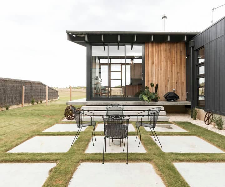 A modern home design with a wooden and black facade features a patio with a table and chairs, surrounded by greenery and stone pathways.