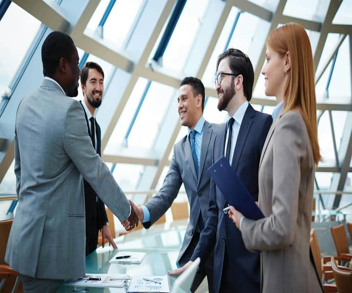 A business meeting in a modern office, featuring professionals shaking hands, discussing, and holding documents at a glass conference table.