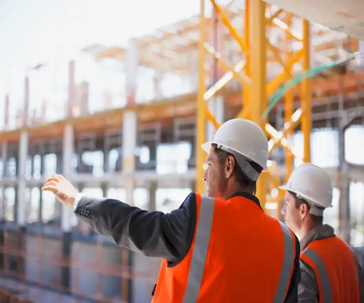 Two construction workers in hard hats and safety vests observe a building site, one gesturing towards the structure to ensure the building regulations