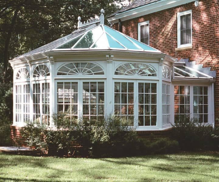 Bright, elegant conservatory attached to a brick house, featuring large windows, a glass roof, and surrounded by lush greenery.