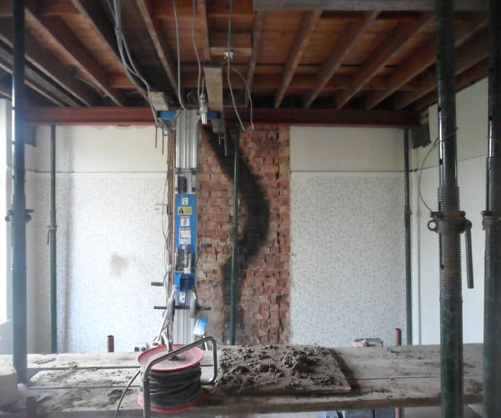 A partially demolished room with exposed brick wall, construction support beams, and tools on a workbench.