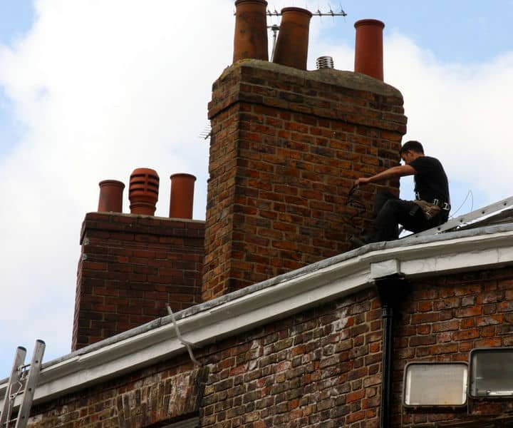 A worker on a rooftop adjusts a chimney over a brick building, surrounded by multiple vent stacks under a cloudy sky.