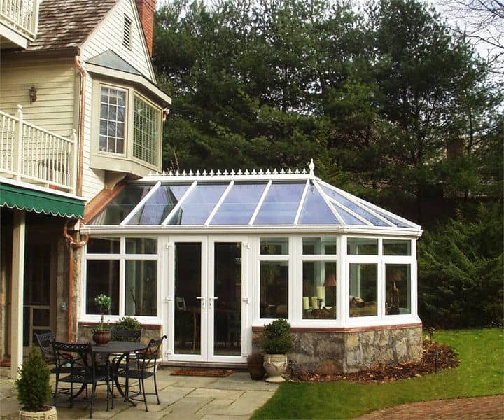 A spacious conservatory style sunroom with white framing, attached to a house, surrounded by greenery.