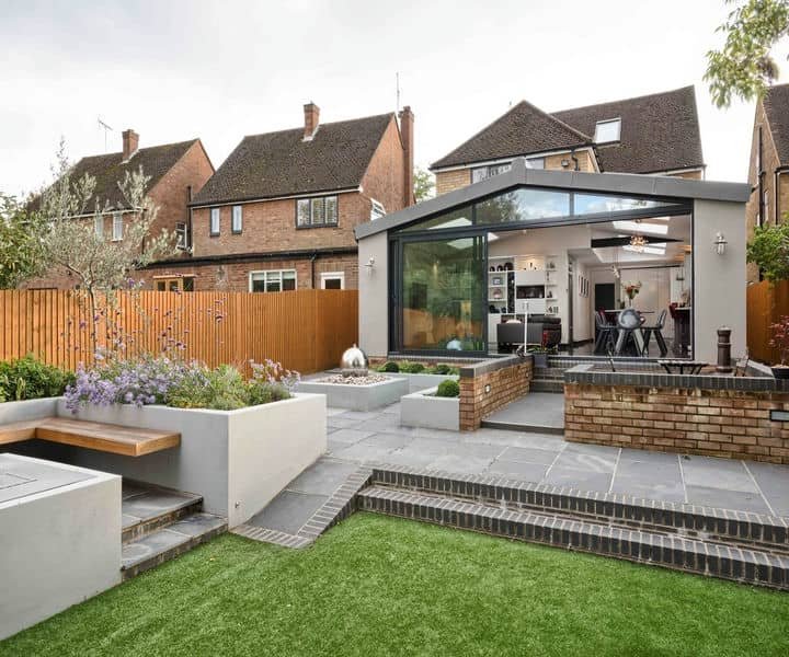 Modern garden with a glass conservatory extension, featuring a lawn, stone patio, raised beds, and a wooden fence, surrounded by brick houses.