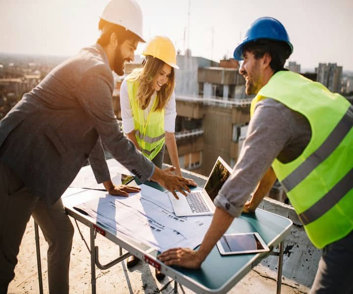 Three professionals in safety vests and hats collaborate over blueprints and a laptop on a rooftop.