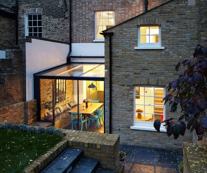Arear extention for a cosy modern kitchen with glass walls, featuring a wooden dining table, colorful chairs, and warm lighting at dusk.