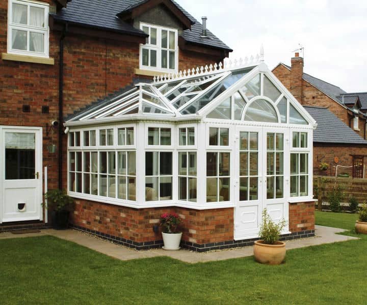 A charming white edwardian conservatory attached to a brick house, featuring large glass windows and a peaked roof, surrounded by a green lawn.
