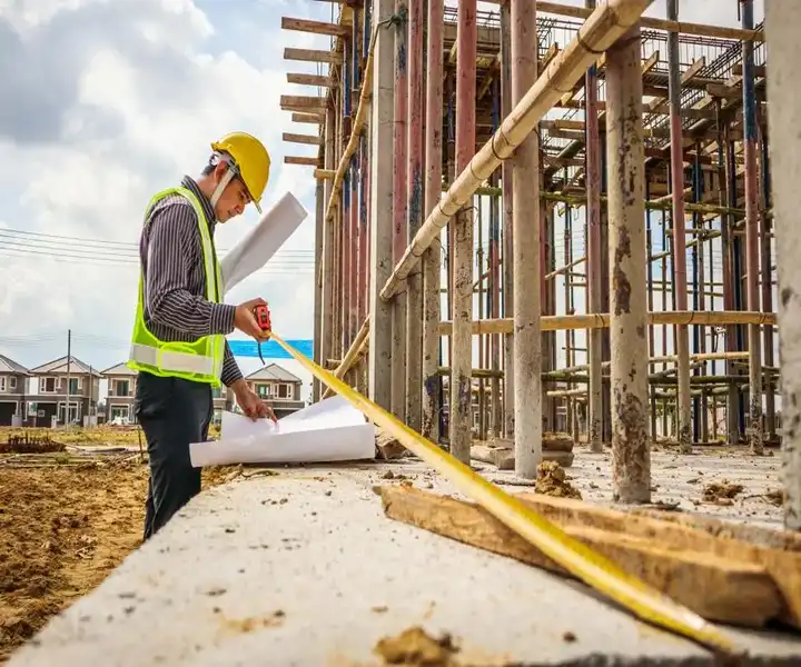 An engineer measures with a tape on a building site, holding blueprints amidst scaffolding and a clear sky.