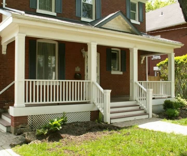 A charming brick house with a white front porch, featuring green shutters, a well-maintained lawn, and lush greenery nearby.