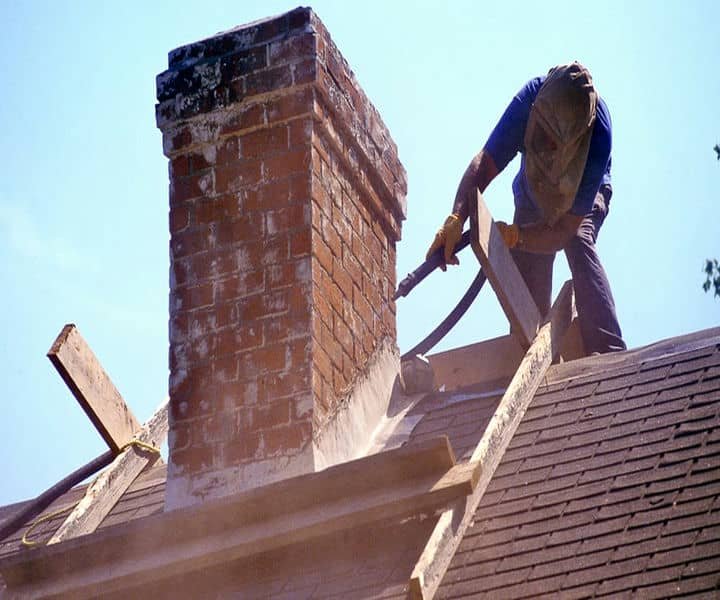 A worker wears protective gear while using a power tool to remove a chimney on a rooftop.