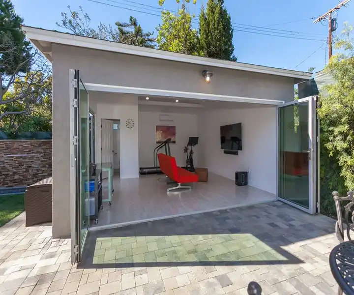 Garage conversion into a Modern studio with glass doors, featuring a fitness space, red chair, and a TV, surrounded by greenery and a stone patio.