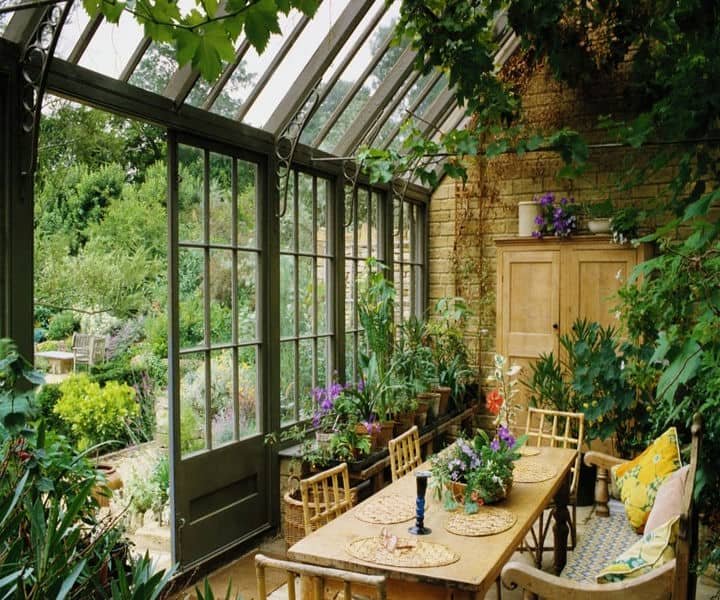 A sunlit garden room with a wooden dining table, surrounded by lush plants and flowers, offering a view of a vibrant garden outside.