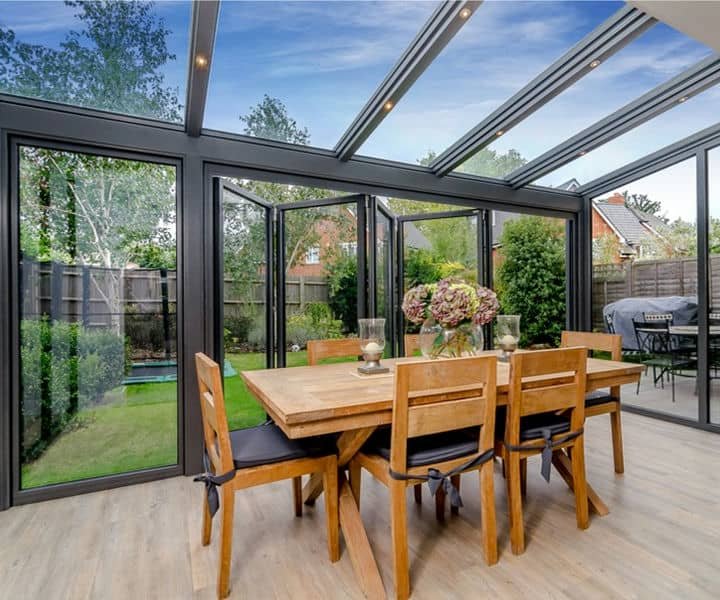 Bright sunroom desined with a dining area, surrounded by glass walls and ceiling, showcasing a lush garden view outside.