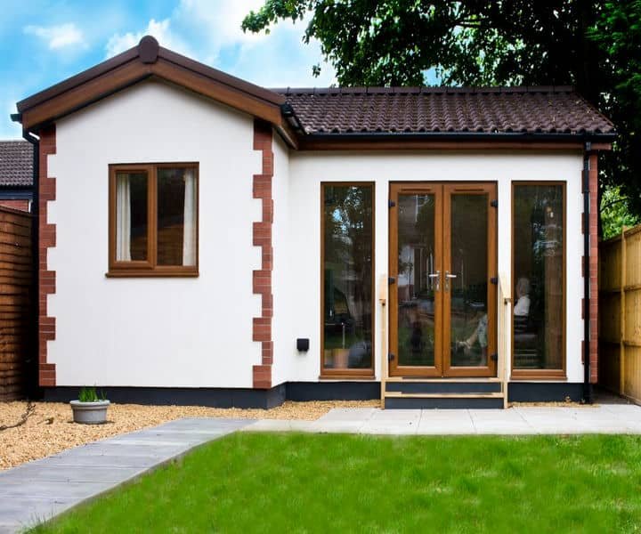 A modern granny annexe with wooden doors, large windows, and a peaceful garden setting under a blue sky.