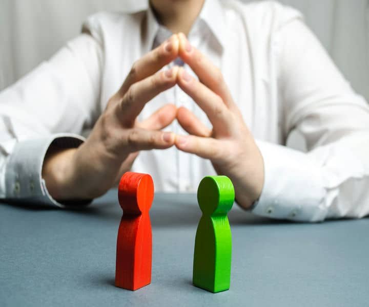 A person in a white shirt sits with hands clasped, contemplating two wooden figures, one red and one green, symbolising choice between two opposition.