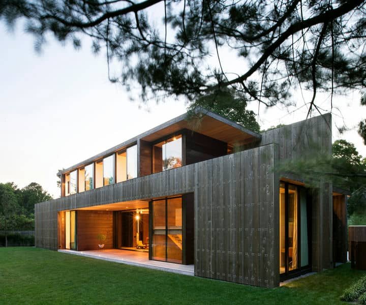 Modern architectural deigned two-story wooden house with large windows, set in a green landscape at dusk, showcasing minimalist architecture and natural surroundings.
