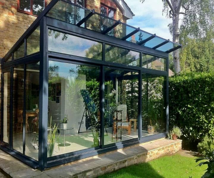 Modern glass sunroom with a black frame, filled with plants and furniture, surrounded by greenery and sunlight.
