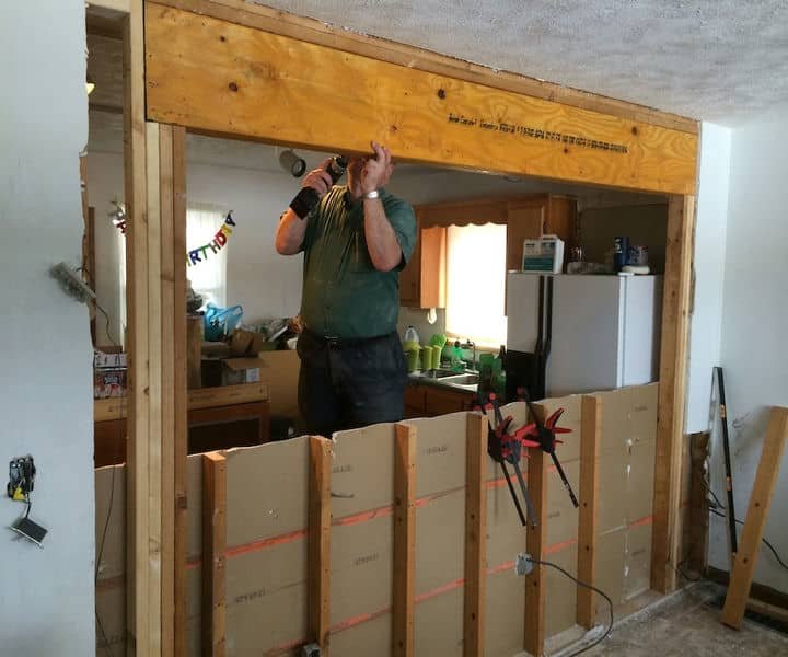 A man installs a wooden beam above a doorway, surrounded by construction materials in a partially renovated kitchen.