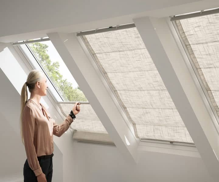 A woman adjusts a sleek, patterned roller shade on a skylight in a modern, bright loft bedroom filled with natural light.