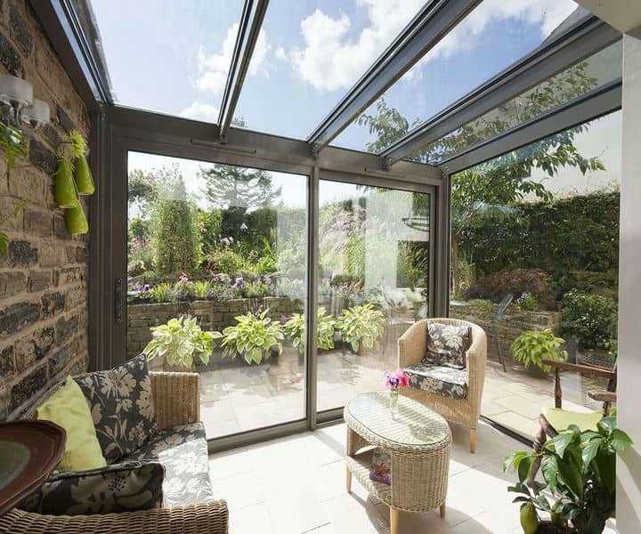 Bright sunroom with glass walls, featuring comfortable seating,, and lush garden views through the large windows.