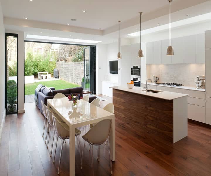 Modern open-plan kitchen and dining area with wooden floors, sleek furniture, and large glass doors leading to a green garden.