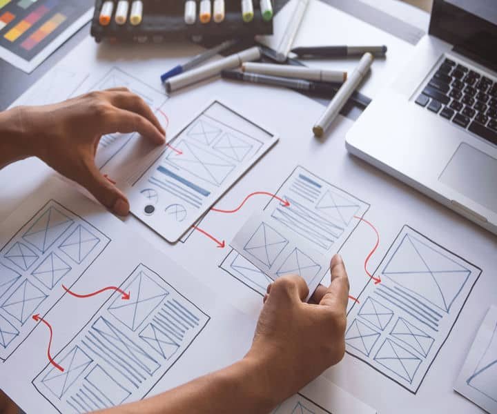 A designer's hands manipulate wireframe sketches, linking them with red arrows, surrounded by markers and a laptop on a table.