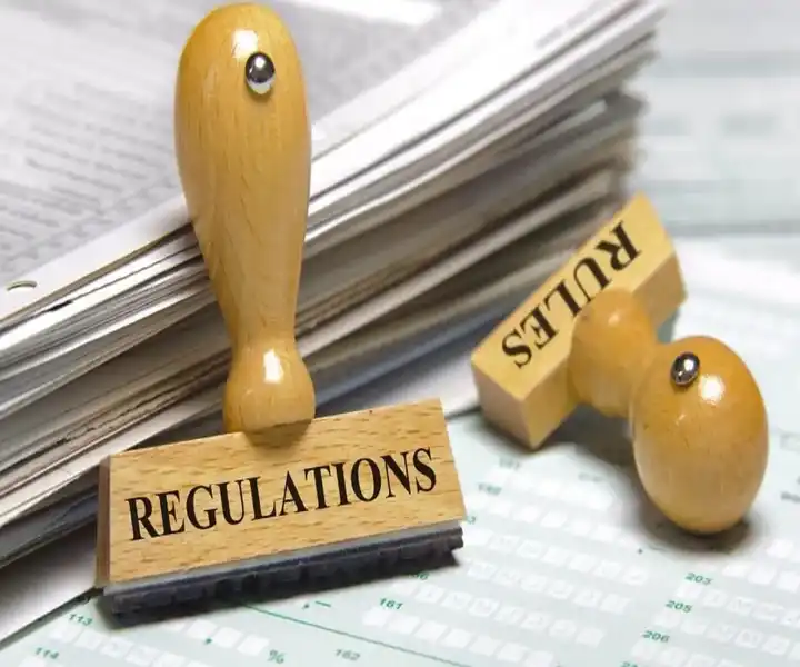 Two rubber stamps labeled "REGULATIONS" and "RULES" resting on a pile of papers, symbolising compliance and legal documentation.