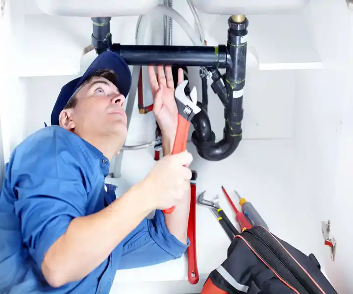 A plumber works under a sink, using a wrench to adjust pipes, with various tools and a toolbox nearby.