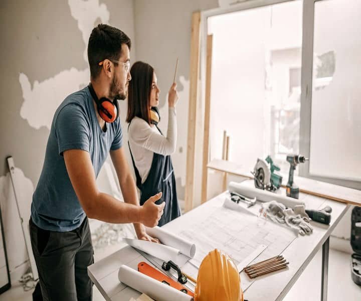 A woman points at a blueprint while a man gestures, surrounded by construction tools and materials in a partially renovated room.