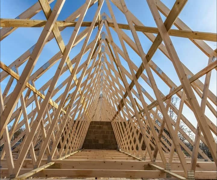 Wooden roof frame under construction, showcasing a geometric pattern of beams against a clear blue sky.