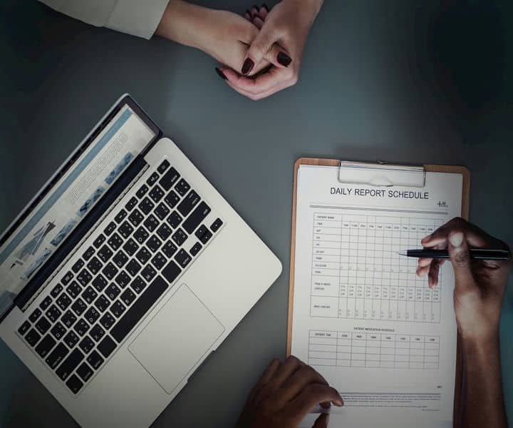 A laptop and a clipboard with a daily report schedule, with two hands engaged in discussion or note-taking.