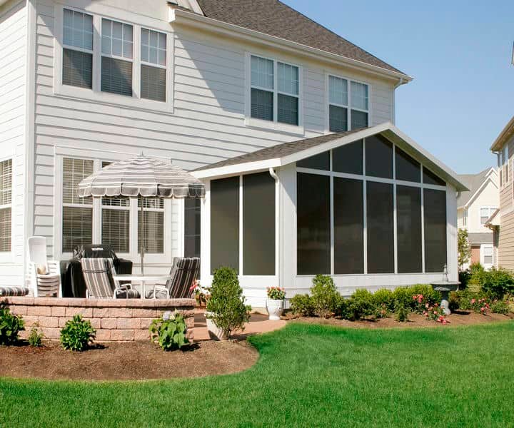A sunny backyard featuring a screened porch, patio with striped umbrella, and landscaped garden beside a two-story home.