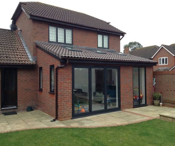 A modern single-story brick house extension with large glass doors opening to a garden, featuring a lawn and patio area.