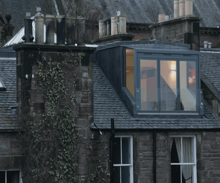 A modern glass-walled loft extension over a historic stone building, featuring chimneys and ivy on the wall.