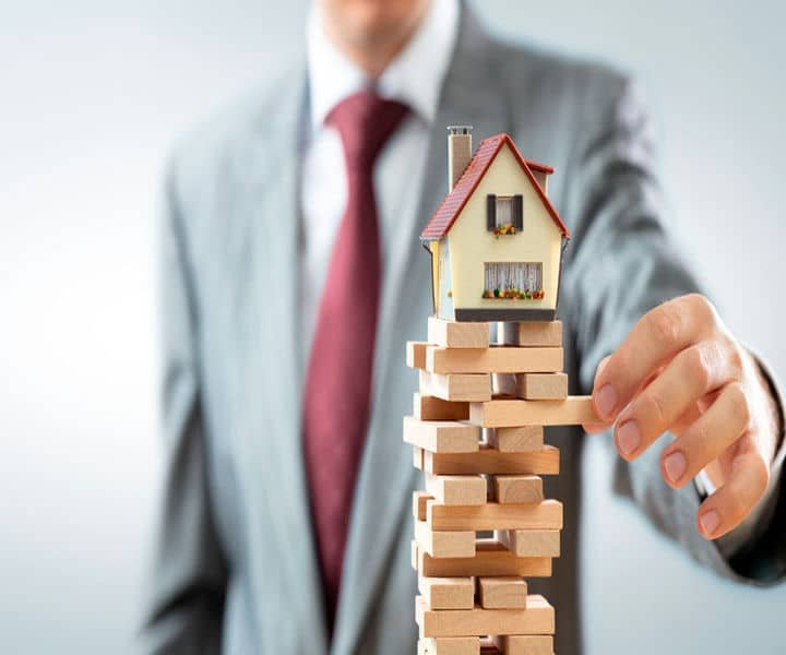 A person in a suit carefully balances a miniature house over a tower of wooden blocks, symbolising risk in real estate.