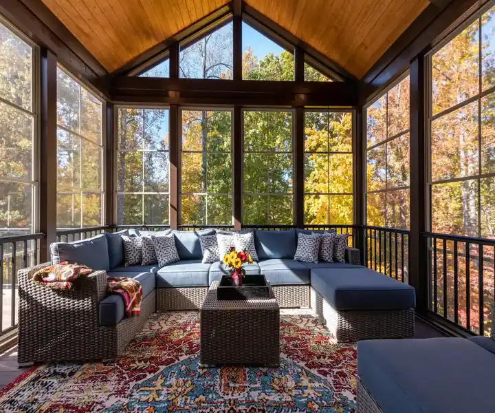 A cosy sunroom featuring a large blue sectional sofa, a decorative rug, and vibrant autumn foliage visible through the windows.