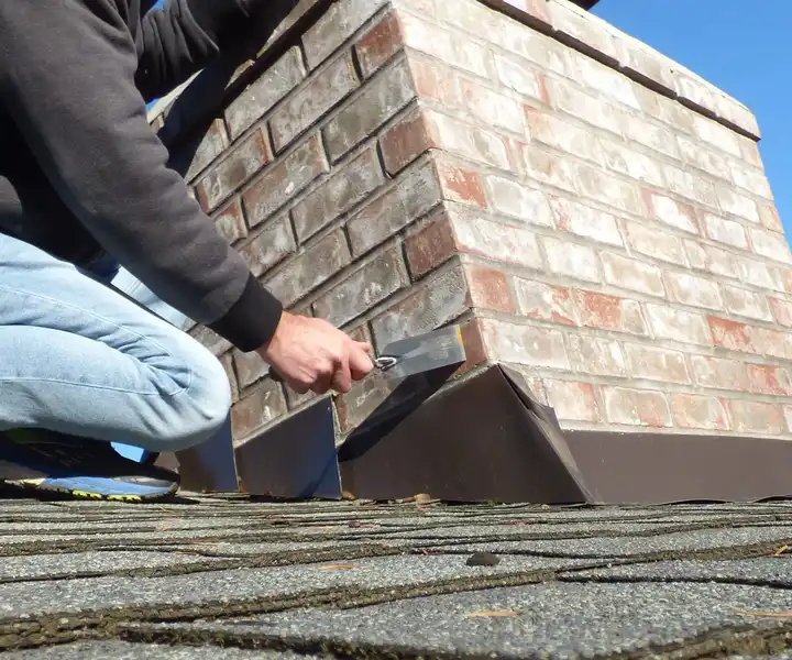 A person repairs a brick chimney on a rooftop using a trowel, with clear blue skies in the background.