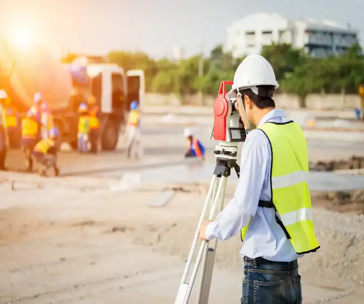 A surveyor with a helmet and reflective vest uses a surveying instrument at a site, with workers and vehicles in the background.