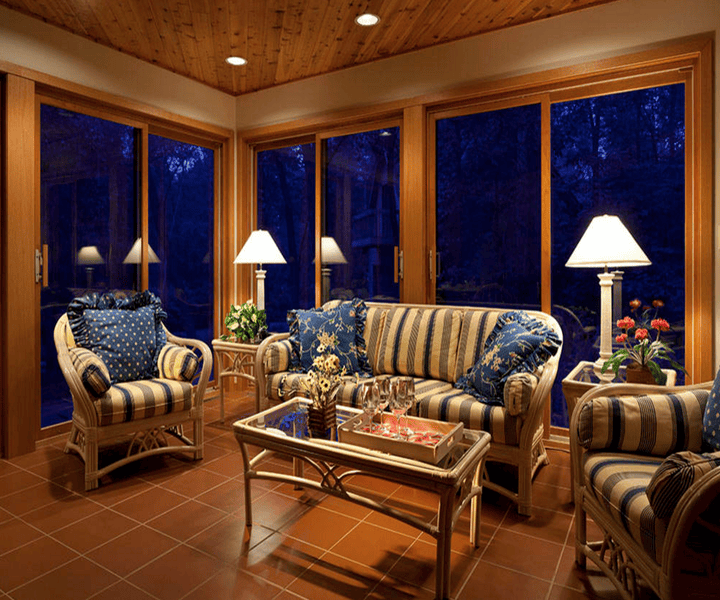 A traditional, cosy sunroom with striped sofas and floral pillows, adorned with lamps, overlooking a darkened outdoor landscape.