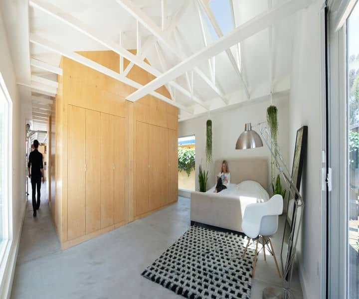 Trending garage conversion into a bright, modern bedroom with wooden cabinetry, open ceiling beams, and a cosy white bed, featuring a woman reading.
