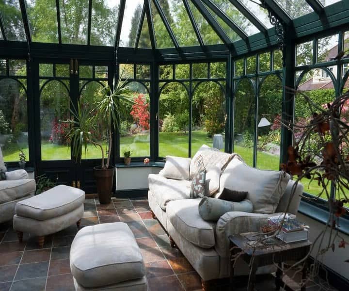A bright sunroom with a plush sofa, , and greenery, surrounded by large glass windows.